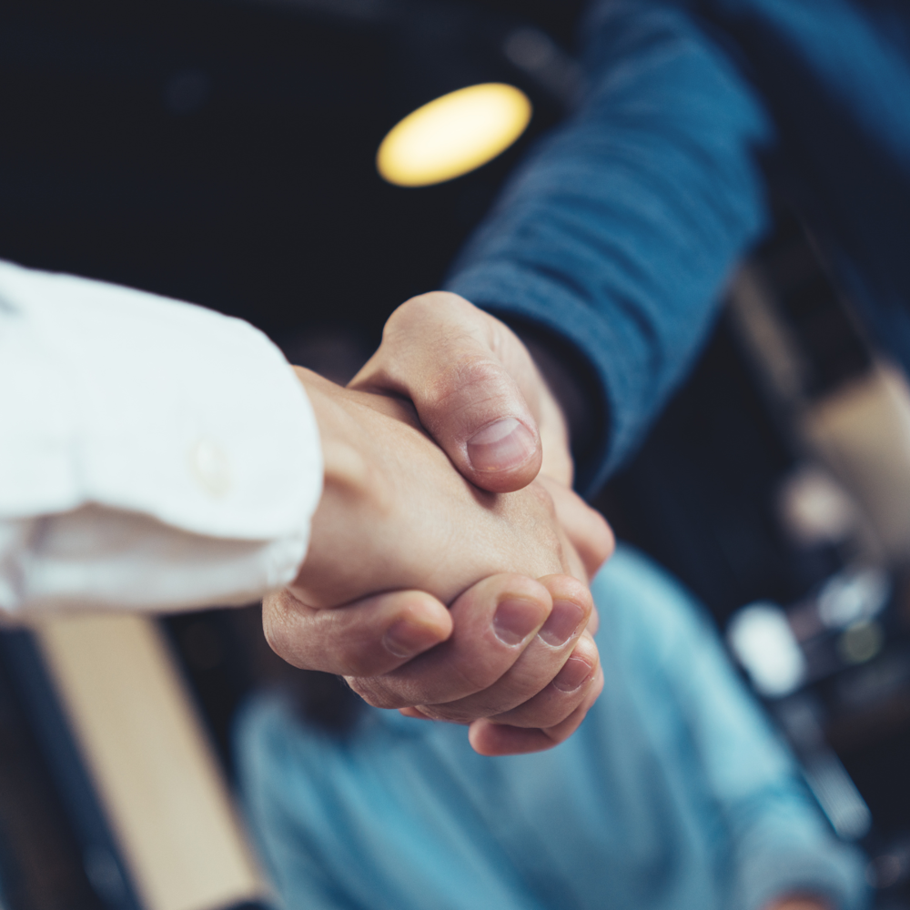 A close-up of a handshake between two people in an informal business setting, with a softly blurred background.