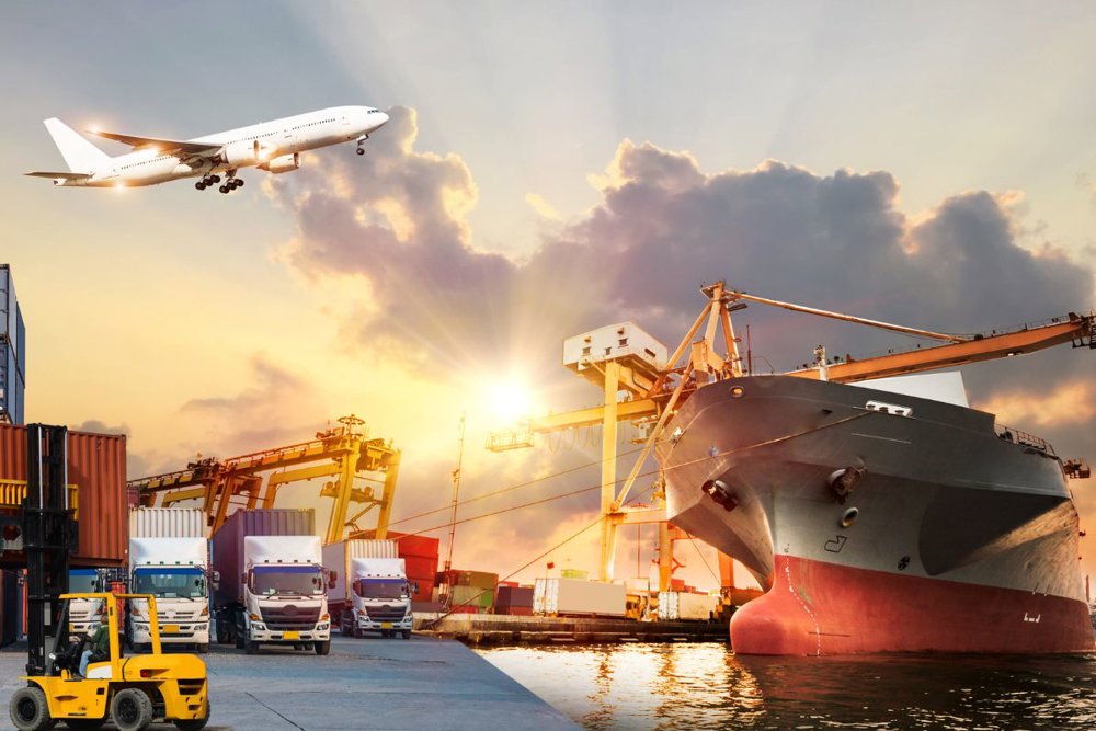 Pristanišče ob sončnem zahodu, kjer je na pomolu privezana velika ladja za prevoz tovora. Ob njej stojijo kontejnerji in več dvigal, ki nakladajo ali razkladajo tovor. Na levi strani je viden viličar pred kupom kontejnerjev, v ozadju pa so parkirani tovorni tovornjaki. Nad prizorom leti potniško letalo.