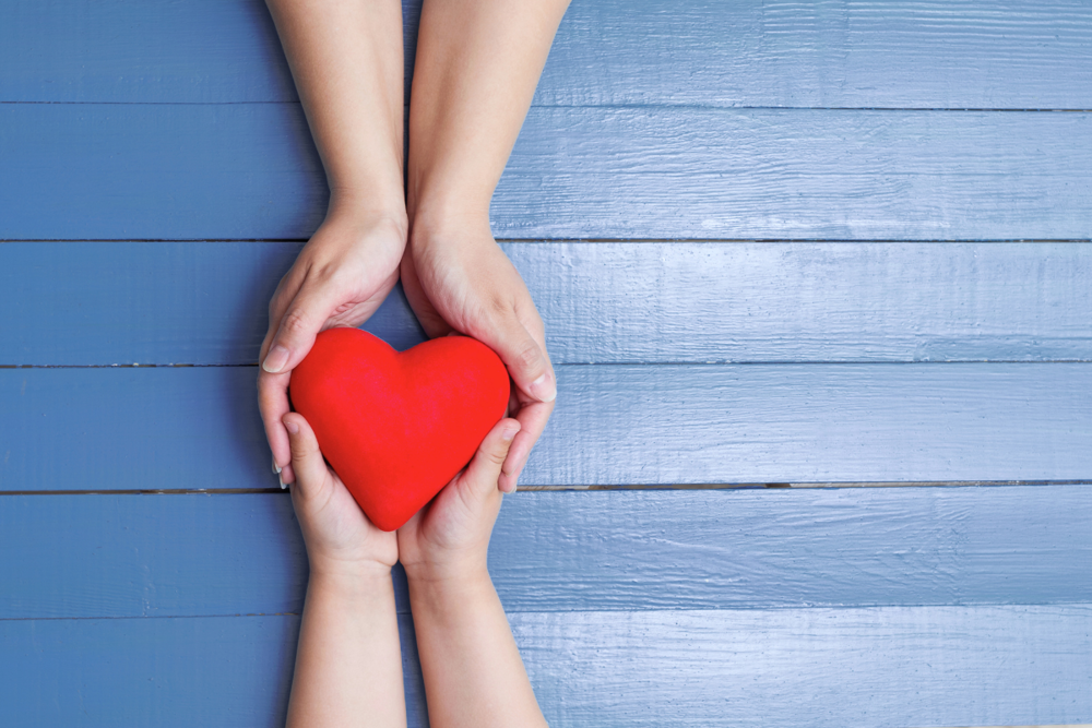 Two hands give a red heart to another pair of hands on a blue wooden background.