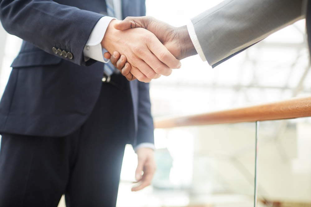 Dve osebi v poslovnih oblekah si segata v roke, prikazan je stisk rok v svetlem poslovnem okolju.