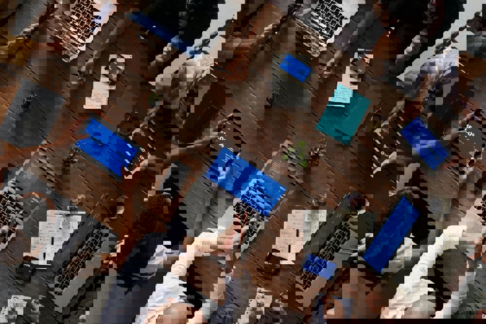 Skupina ljudi sedi okoli lesene mize in dela na prenosnikih, tablicah in telefonih. Na mizi so tudi zapiski, grafikoni, zvezki in skodelice s pijačo, kar prikazuje timsko analizo ali sodelovanje.