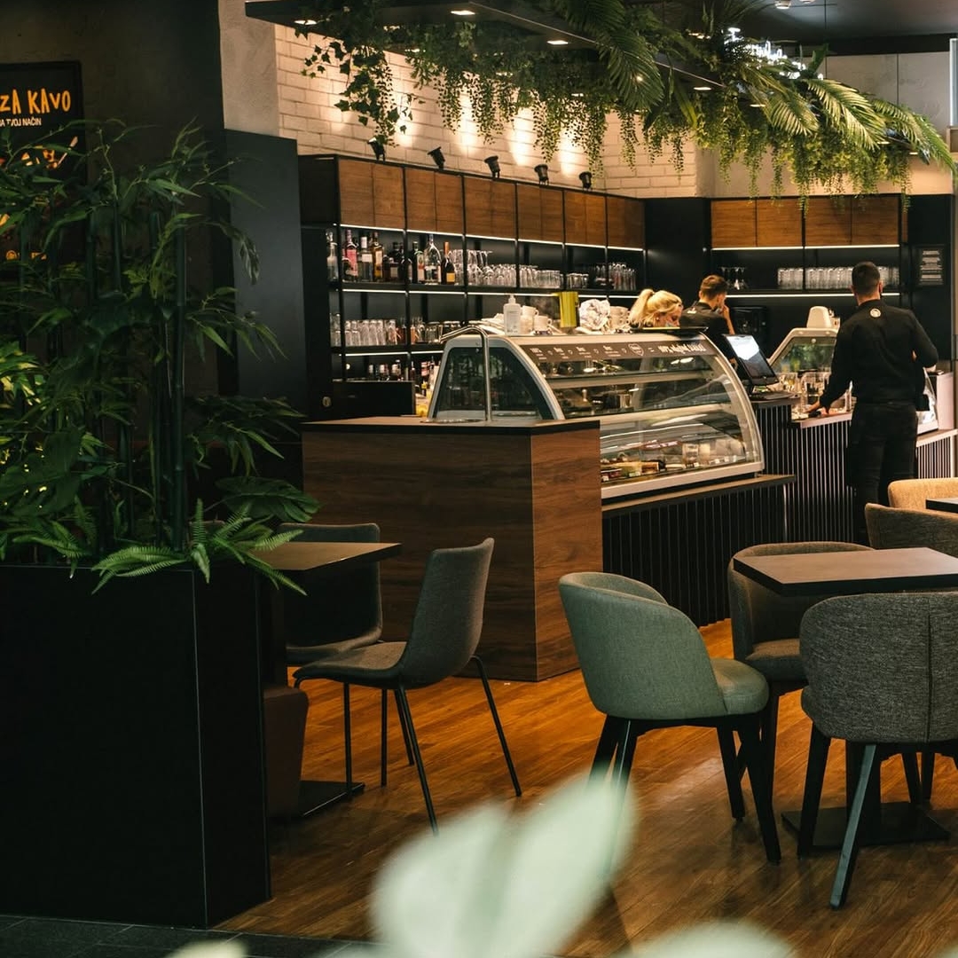 Interior of a modern café with wooden floors, tables and upholstered chairs, a dessert display counter, and hanging green plants above the service area.