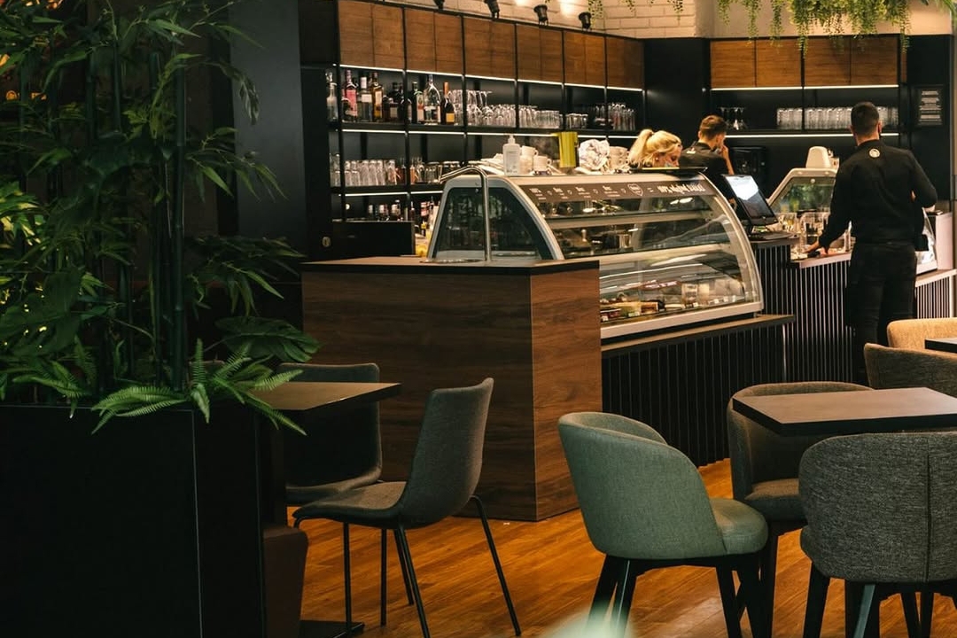Interior of a modern café with wooden floors, tables and upholstered chairs, a dessert display counter, and hanging green plants above the service area.