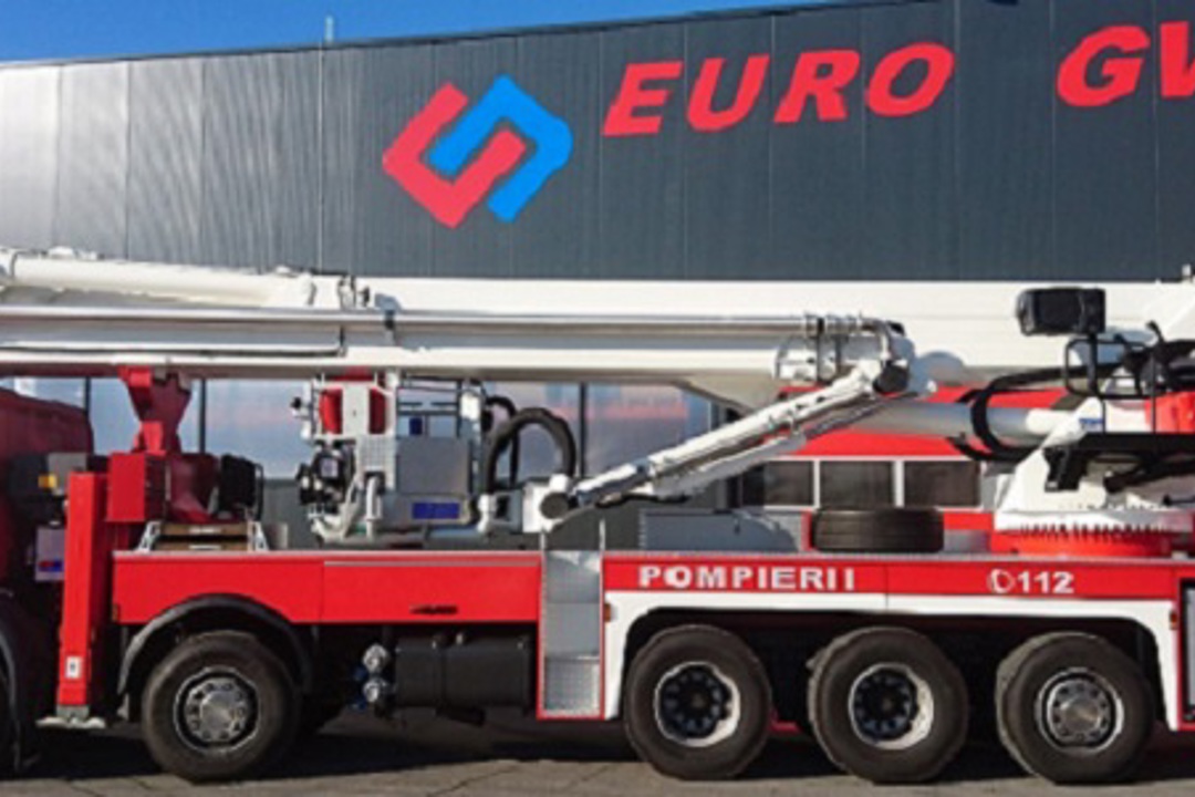 Na parkirani površini pred industrijsko stavbo je specialno gasilsko vozilo z dolgo dvižno roko in opremo za intervencije.