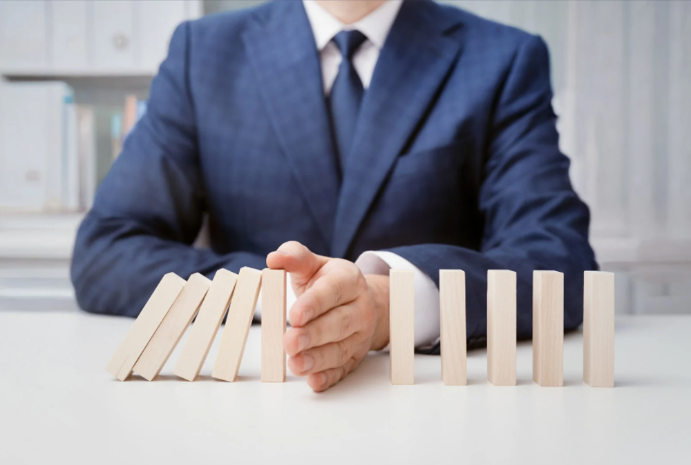 Oseba v temnem poslovnem suknjiču z dlanjo ustavlja padajoče lesene domine na beli mizi.