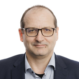 Oseba v temnem suknjiču in svetli srajci pred belim ozadjem.