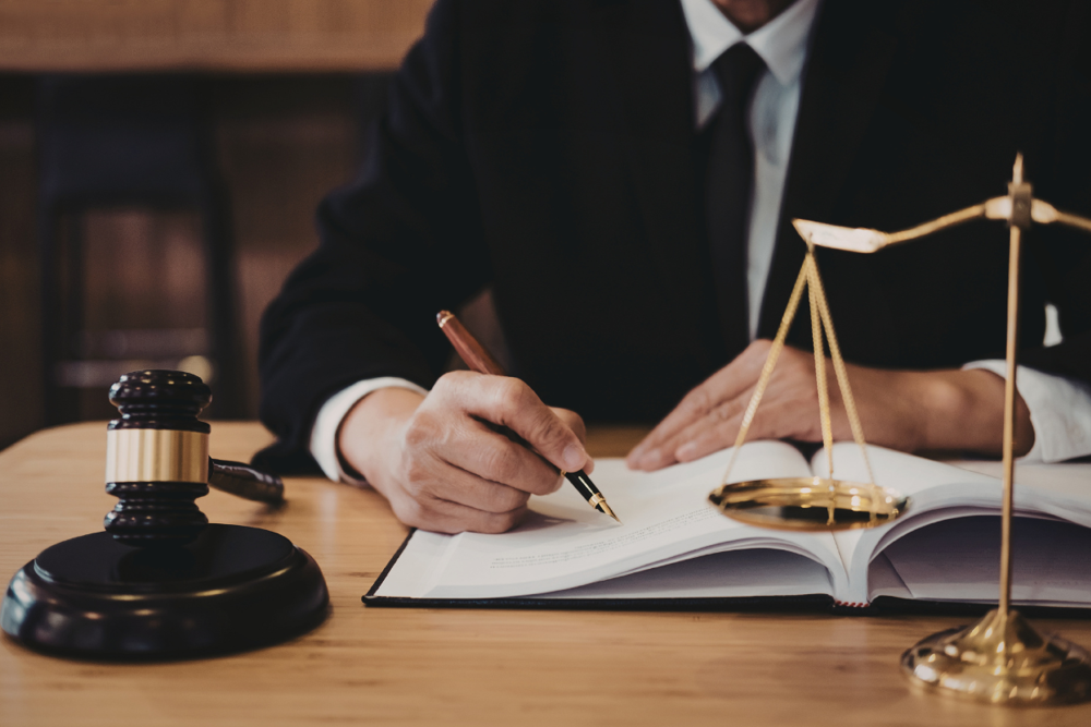 Oseba v poslovni obleki piše v odprto knjigo na mizi, ob njej pa sta sodniški kladivce in tehtnica pravice.