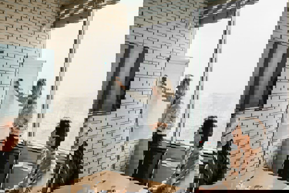 Oseba stoji ob prezentacijski tabli in piše na papir, medtem ko dve osebi za mizo spremljata predstavitev.