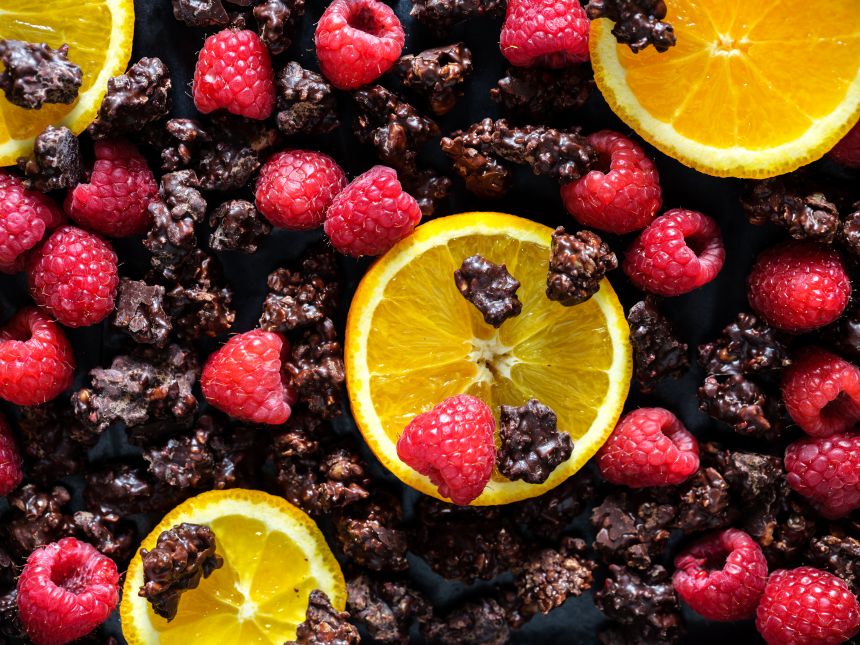 Slices of orange, fresh raspberries, and clusters of dark chocolate are arranged on a dark surface.
