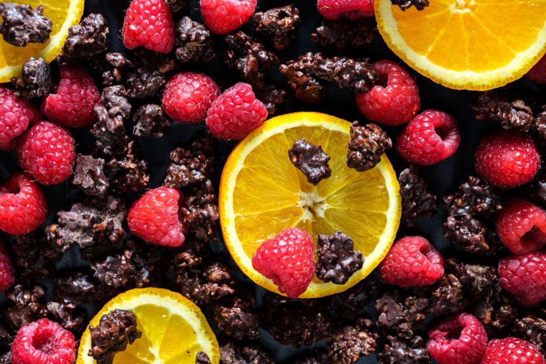 Slices of orange, fresh raspberries, and clusters of dark chocolate are arranged on a dark surface.