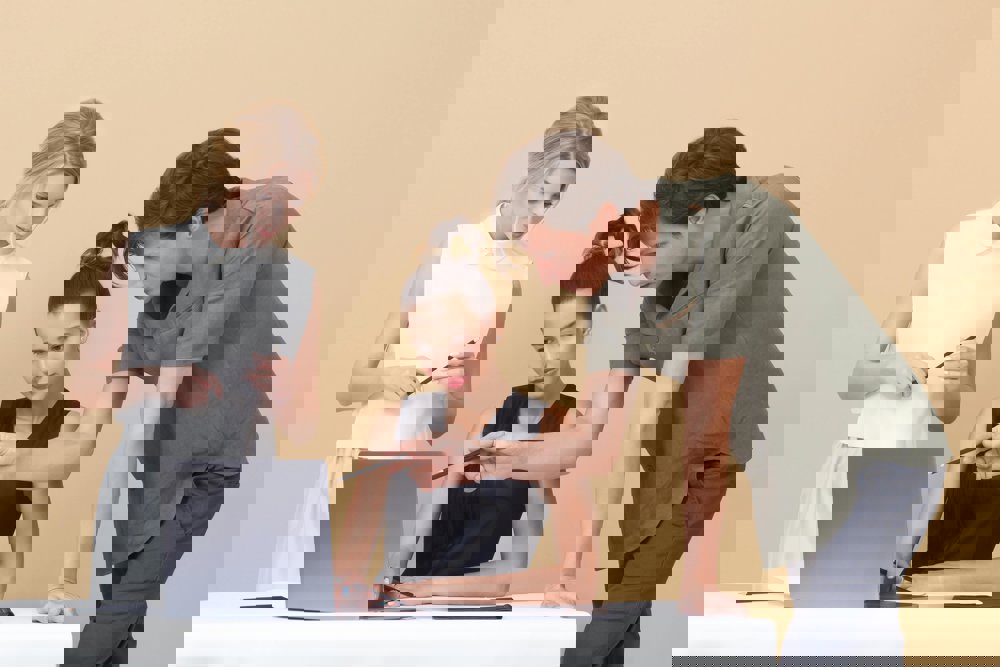 Tri osebe sodelujejo pri delu ob prenosniku; ena sedi za mizo, druga stoji ob njej in kaže na zaslon, tretja pa stoji ob strani in drži pisalo.