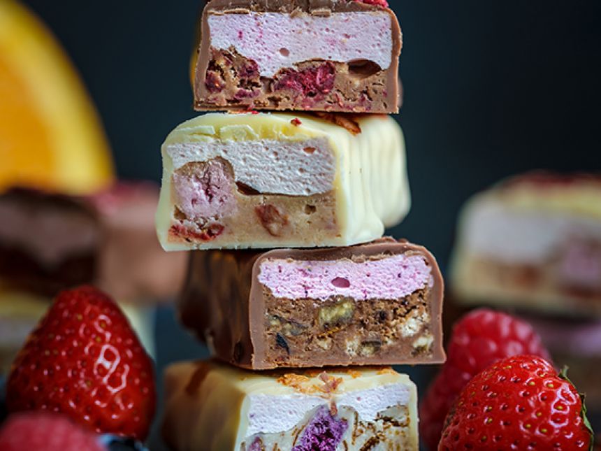 A stack of sliced chocolate bars filled with pink and white fluffy layers and pieces of berries is arranged with fresh strawberries around it.