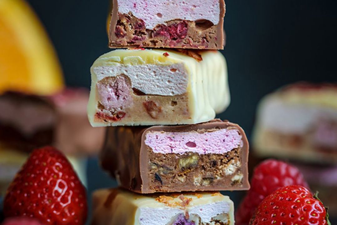 A stack of sliced chocolate bars filled with pink and white fluffy layers and pieces of berries is arranged with fresh strawberries around it.