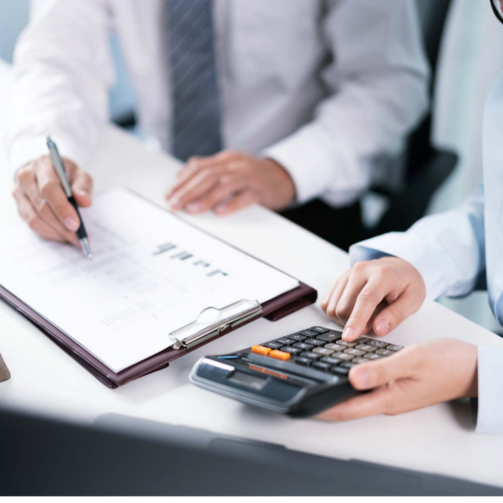 Dve osebi sedita za mizo in pregledujeta finančne dokumente; ena kaže na podatke na papirju, druga pa tipka po kalkulatorju.