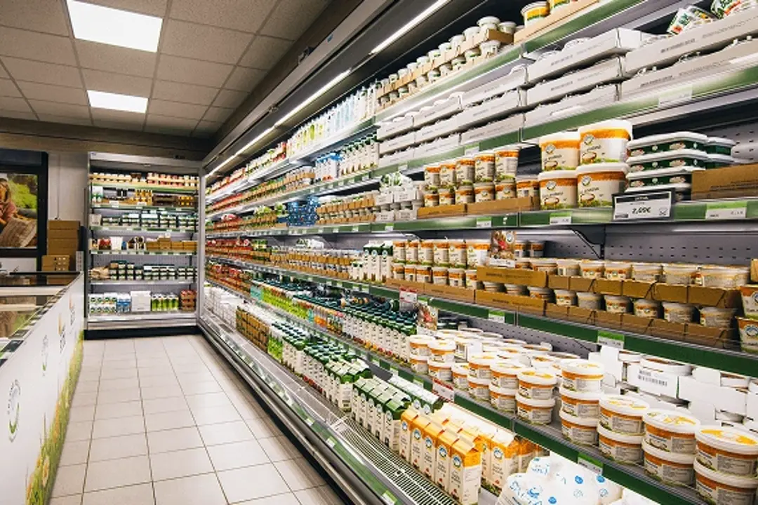 Hladilni regal v trgovini, poln mlečnih izdelkov in embalaž na policah, osvetljen z zgornjimi lučmi in z jasnim pogledom skozi celoten prodajni hodnik.