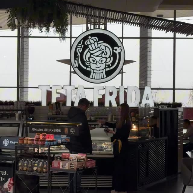 Interior of the Teta Frida shop with an illuminated logo above the counter, where two people stand near the dessert display.