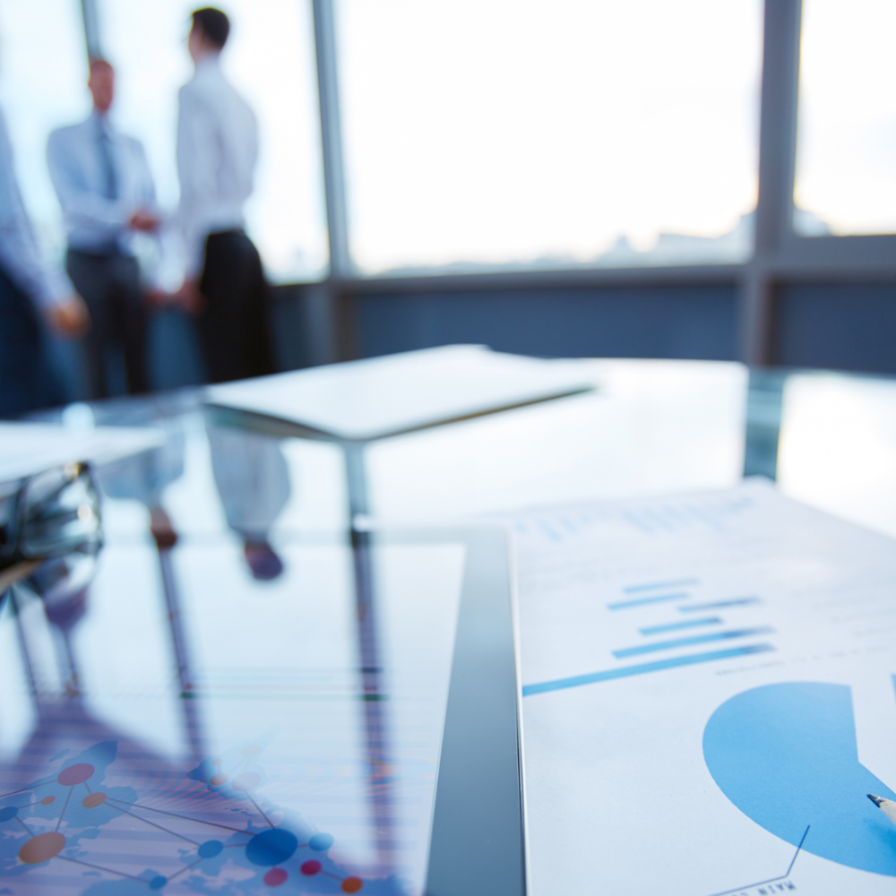 Documents with charts lie on a glass conference table, while three people are visible in soft focus in the background, engaged in conversation in a business setting.